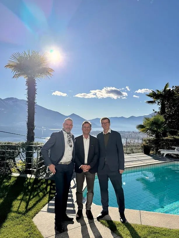 Drei Männer in Anzügen stehen an einem Pool; dahinter eine Palme, ein See und eine Bergkette