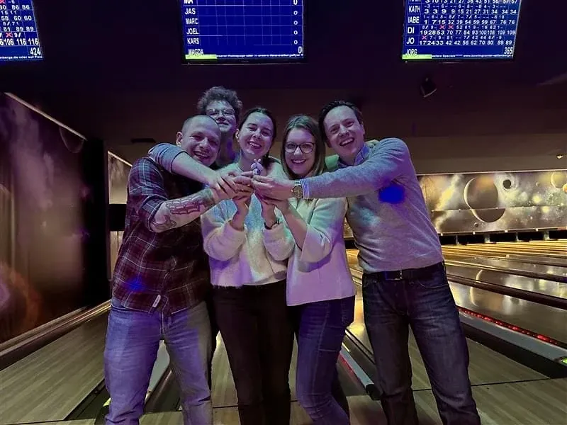 Fünf Personen legen in einer Bowlingbahn lachend die Hände übereinander, darüber die Anzeigetafeln
