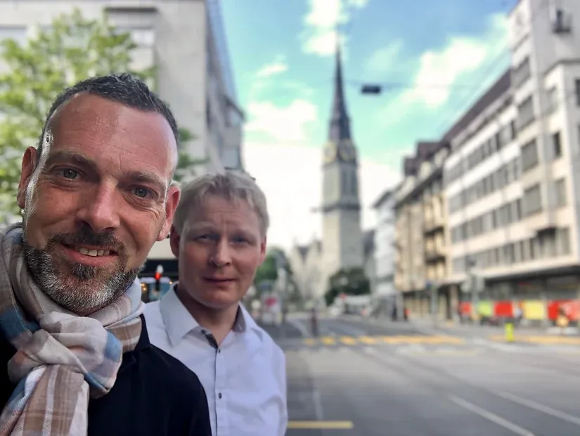 Selfie von zwei Männern auf einer Straße, im Hintergrund ein hoher Kirchturm mit Uhr