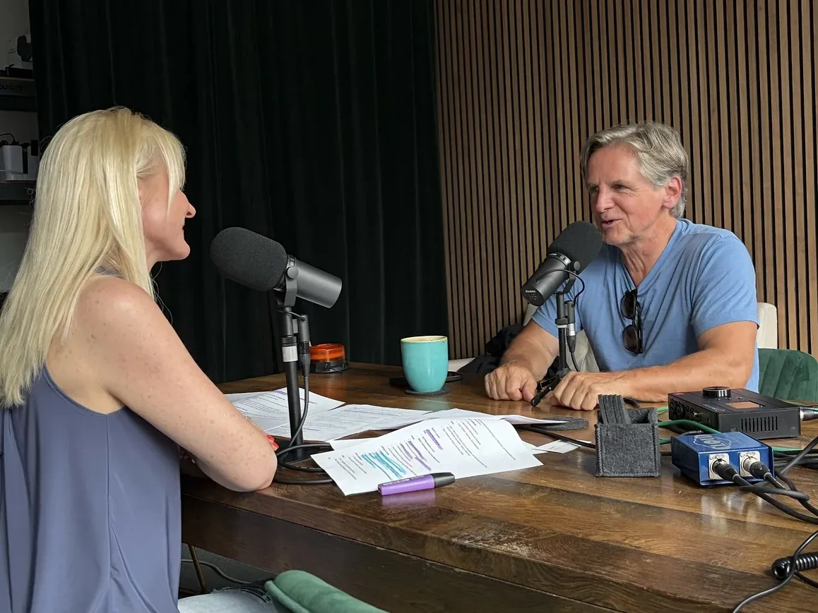 Zwei Personen sitzen sich an einem Holztisch gegenüber, vor jeder ein Podcast-Mikrofon, dazwischen beschriebene Manuskriptseiten