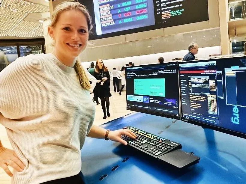 Eine Frau steht lächelnd an einem Bloomberg-Terminal, davor zwei Bildschirme mit Kursdaten