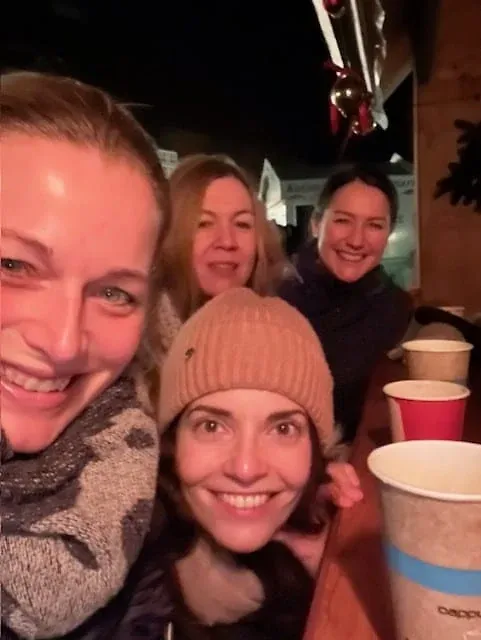 Selfie von vier Frauen mit Bechern in der Hand an einem abendlichen Weihnachtsmarktstand