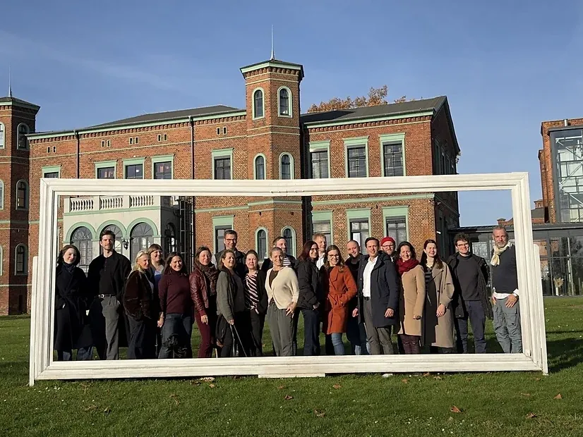 Das Team steht in einem übergroßen weißen Bilderrahmen auf einer Wiese vor einem Backsteingebäude