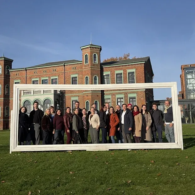 Das Team steht in einem übergroßen weißen Bilderrahmen auf einer Wiese vor einem Backsteingebäude