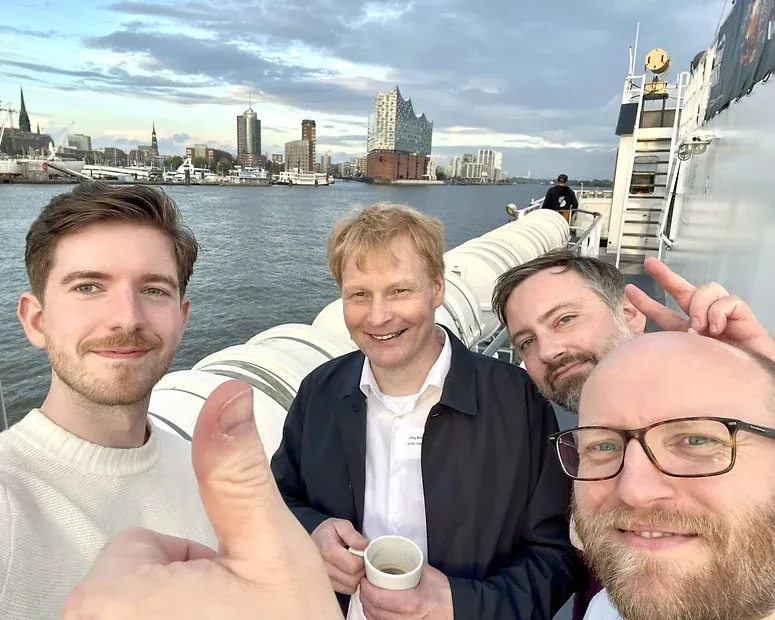Selfie von vier Männern an Bord einer Hafenfähre, dahinter die Elbphilharmonie