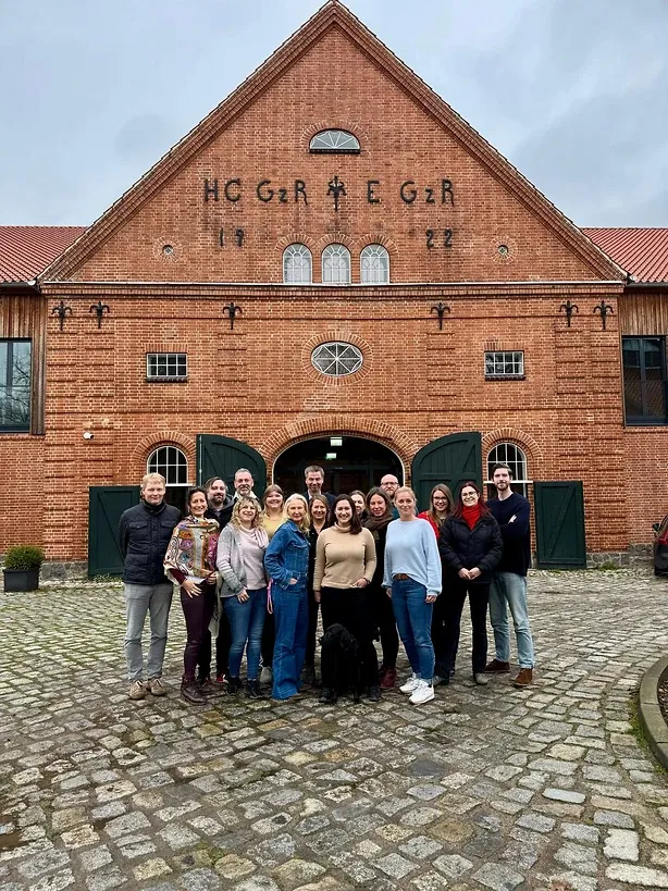 Das Team auf Kopfsteinpflaster vor dem Giebel eines Backsteinhauses mit Ziersteinen im Mauerwerk
