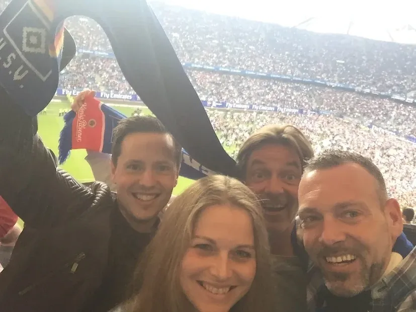 Selfie von vier Personen auf der Tribüne eines vollbesetzten Fußballstadions, darüber ein hochgehaltener Vereinsschal