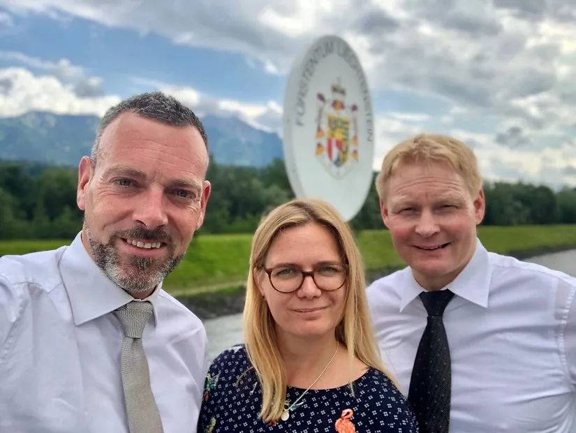 Selfie von drei Personen in Hemd und Krawatte vor dem Wappenschild „Fürstentum Liechtenstein", dahinter ein Fluss und eine Bergkette