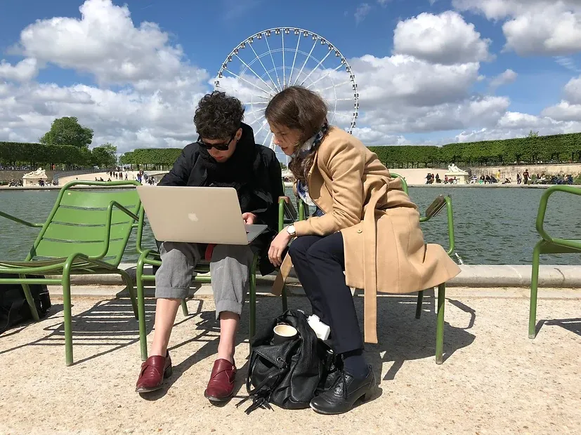 Zwei Personen sitzen mit einem aufgeklappten Notebook auf grünen Gartenstühlen am Wasserbecken eines Parks, dahinter ein Riesenrad