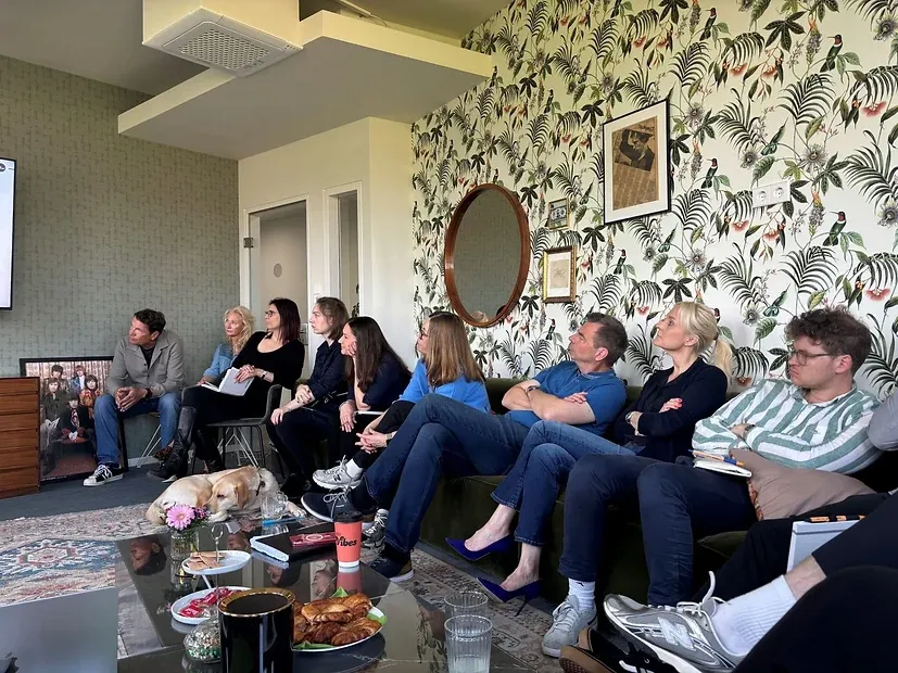 Eine Runde sitzt auf Sofa und Stühlen vor einer Wand mit Blätter-Tapete und blickt zur Leinwand; vorn liegen zwei Hunde
