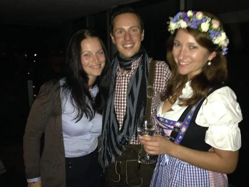 Drei Personen bei einer Abendveranstaltung, zwei davon in Tracht — Lederhose und Dirndl mit Blumenkranz