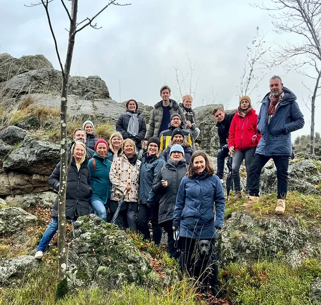 Eine große Gruppe in Outdoorjacken steht mit einem Hund auf einem Felsen in herbstlicher Landschaft