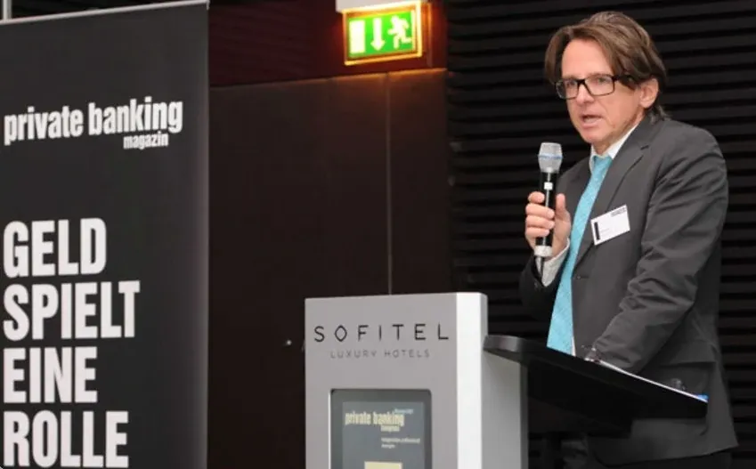 Ein Mann mit Brille spricht ins Mikrofon neben einem Rednerpult; links ein Roll-up des private banking magazins mit der Zeile „Geld spielt eine Rolle“