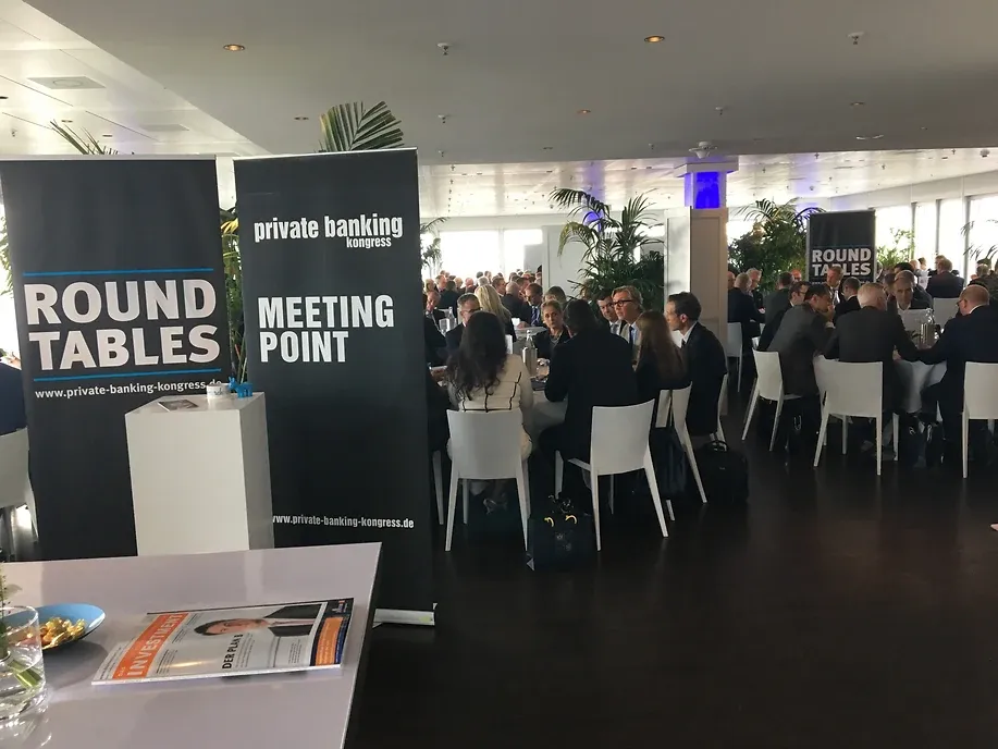 Kongressgäste an runden Tischen in einem hellen Raum mit Fensterfront, davor zwei Roll-ups mit den Aufschriften „Round Tables“ und „Meeting Point“
