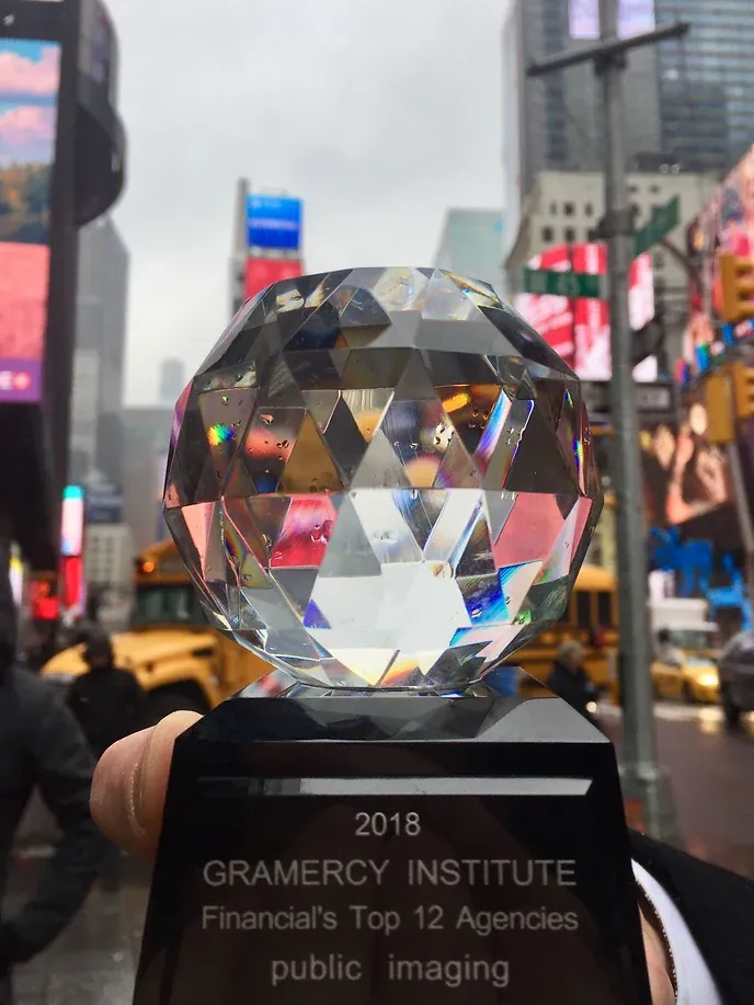 Eine Hand hält eine facettierte Glaskugel auf schwarzem Sockel in die Höhe; im Hintergrund die Leuchtreklamen des Times Square. Der Sockel ist beschriftet mit „2018 Gramercy Institute — Financial’s Top 12 Agencies — public imaging“