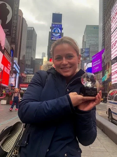 Eine Frau in dunkler Winterjacke hält lachend die Glastrophäe in die Kamera, hinter ihr der Times Square