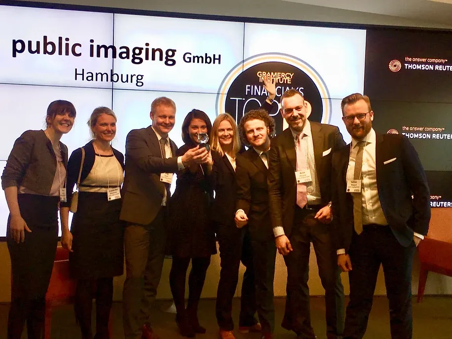 Acht Personen stehen vor einer Videowand, auf der „public imaging GmbH Hamburg“ und das Emblem des Gramercy Institute zu sehen sind; eine hält die Glastrophäe hoch