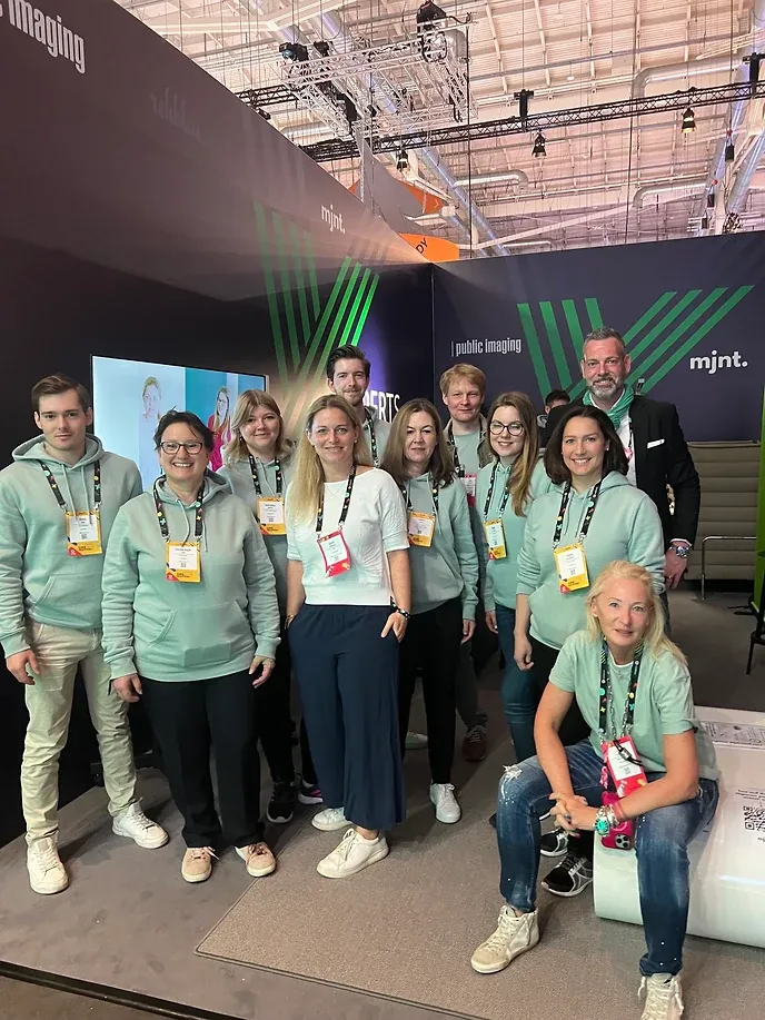 Elf Personen in mintfarbenen Hoodies auf einem Messestand, im Hintergrund eine Wand mit den Logos von public imaging und mjnt