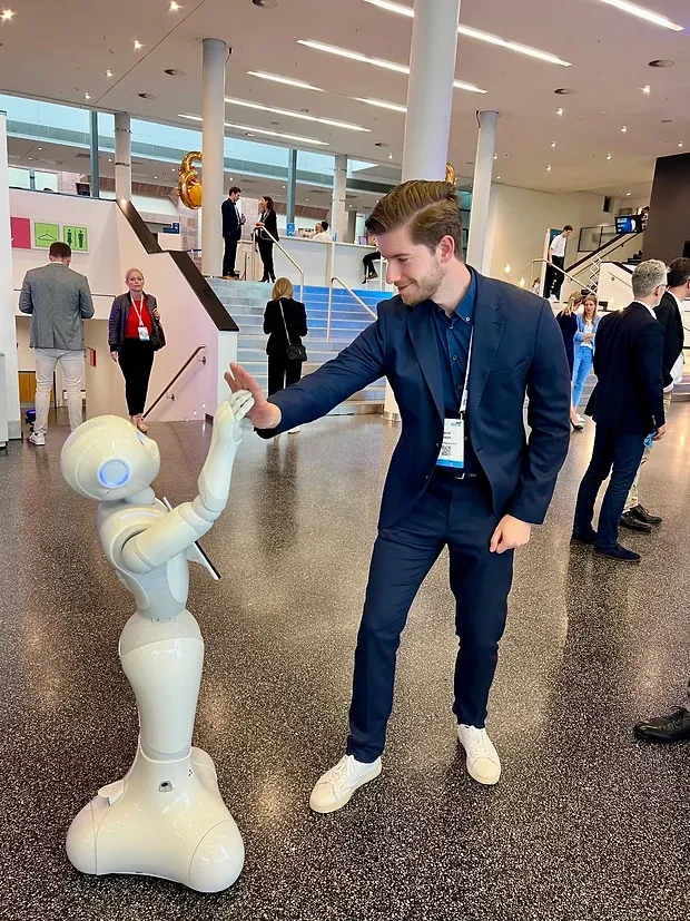 Ein Mann im Anzug schlägt in einem Foyer ein, ein humanoider Roboter hebt ihm die Hand entgegen