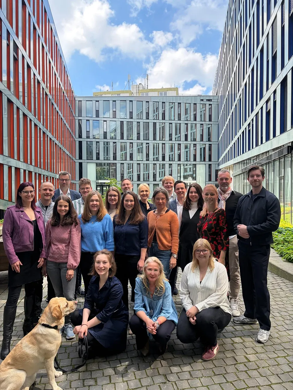 Gruppenbild von rund zwanzig Personen in einem Innenhof zwischen Bürogebäuden, vorne sitzt ein heller Hund