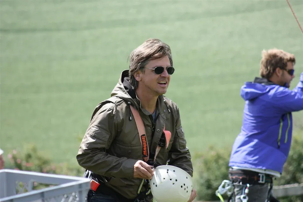 Ein Mann in Kletterjacke mit Sonnenbrille hält einen weißen Helm in der Hand, hinter ihm eine Wiese und eine zweite Person am Sicherungsseil