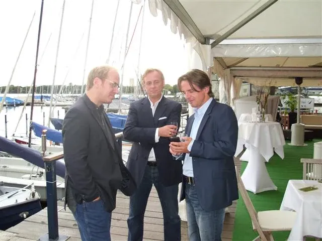 Drei Männer im Sakko stehen mit Gläsern auf einem Steg vor einem Segelhafen, daneben Stehtische unter einem Zeltdach