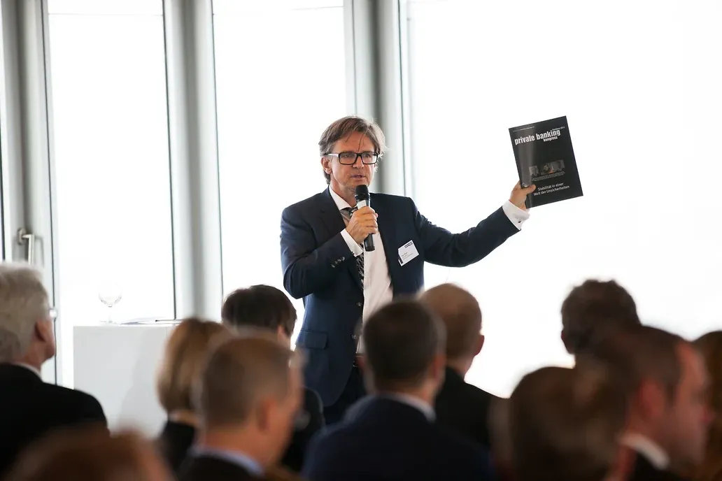 Ein Mann im blauen Anzug spricht vor Publikum ins Mikrofon und hält ein schwarzes Programmheft hoch: „private banking kongress Hamburg 2014 — Stabilität in einer Welt der Unsicherheiten“