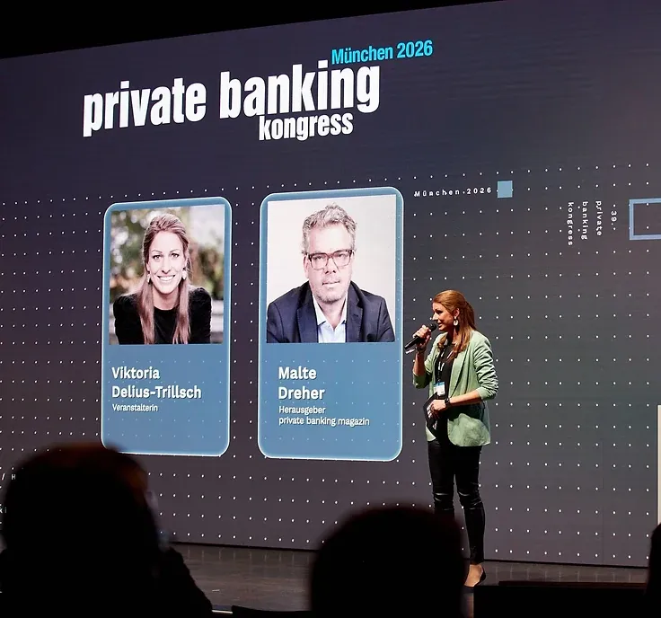 Eine Frau mit Mikrofon steht auf einer Bühne vor einer großen Leinwand mit dem Schriftzug „private banking kongress München 2026“ und zwei Porträtkacheln