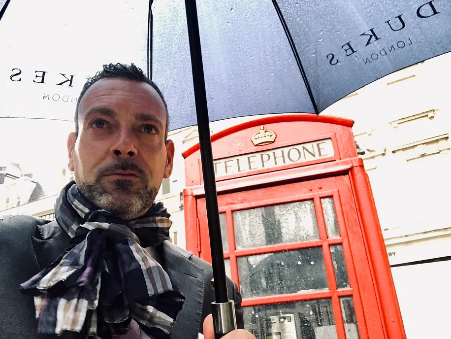 Selfie eines Mannes unter einem Regenschirm mit dem Aufdruck „Dukes London“, dahinter eine rote britische Telefonzelle im Regen