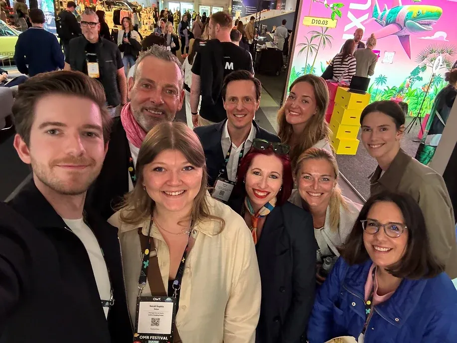 Selfie von neun Personen mit Festivalausweisen in einer Messehalle, dahinter eine pinke Wand mit einer Palmen- und Hai-Illustration