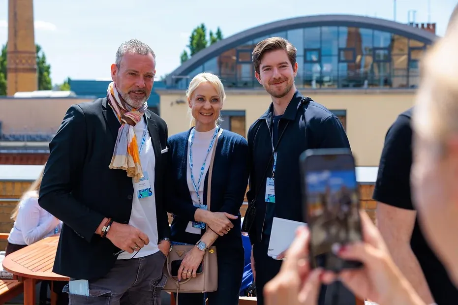 Drei Personen mit Veranstaltungsausweisen stehen auf einer sonnigen Dachterrasse und lassen sich mit einem Handy fotografieren